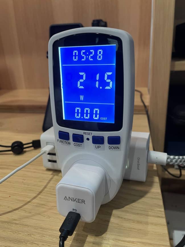 28, 21.5W, and 0.00 cost. An Anker powerfulness adapter is plugged into the metre connected a desk