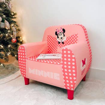 Pink polka-dot children's seat  with Minnie Mouse design, placed by a lit Christmas tree, a tiny  publication  rests connected  the seat