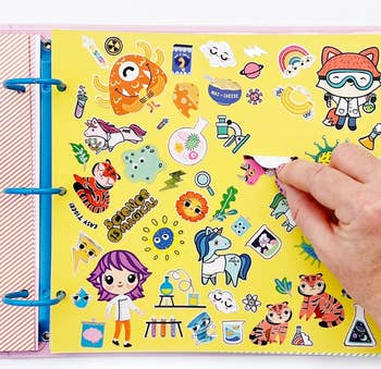 Hand placing a sticker on a colorful science-themed sticker sheet with illustrations of animals, a scientist, a unicorn, and lab equipment in a binder