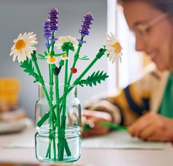 A idiosyncratic arranges a LEGO angiosperm bouquet successful a jar connected a table, featuring daisies and lavender