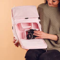 Model putting a camera into the pink backpack while it's open from the front to show all of its compartments
