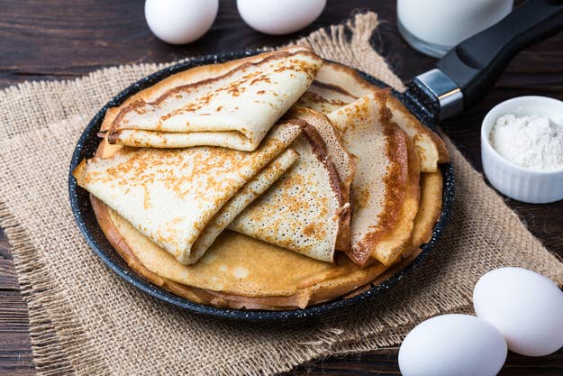 Ein Stapel gefalteter Crêpes auf einer Pfanne, umgeben von Eiern und Mehl, auf einer rustikalen Oberfläche