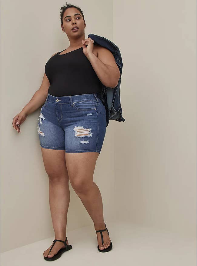 plus-size model wearing medium-wash denim shorts with black strappy sandals and a black top