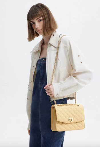 Model in a denim jumpsuit and white jacket, carrying a quilted shoulder bag