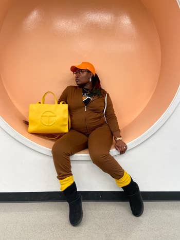 Person posing with a bright yellow tote bag, brown outfit, and black boots against a circular backdrop