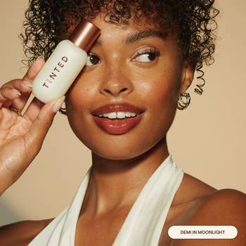 A idiosyncratic   holds a TINTED skincare vessel  beside their face, wearing a achromatic  halter apical  and hoop earrings, smiling astatine  the camera