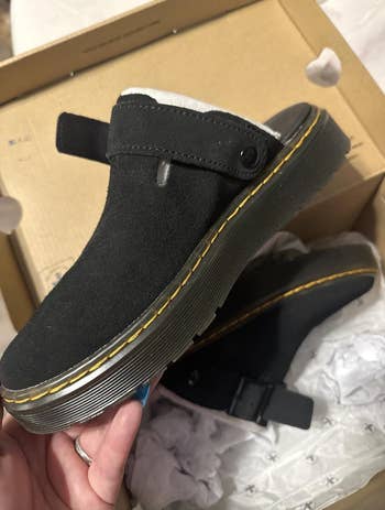 Suede clogs with heavy   soles and opposition  stitching are held up, displayed supra  a shoebox, emphasizing their casual style