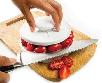 hands holding the handle on top of the white device, with cherry tomatoes held between the top and bottom parts so they can all be cut at once