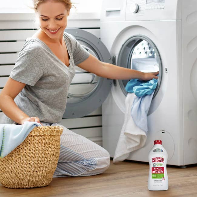 Person happily doing laundry next to a washing machine, holding a basket. A bottle of