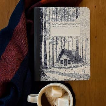 the notebook with a sketch of a house on the cover sitting next to a cup of cocoa  