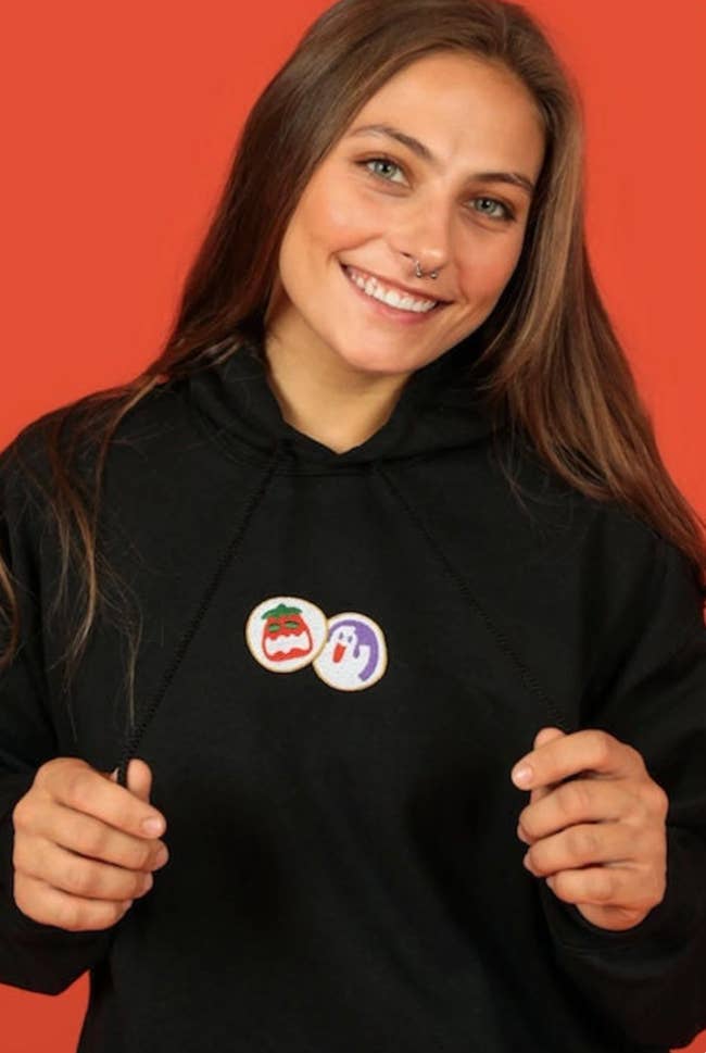 Model in a black drawstring hoodie with two sugar cookies, a jack o lantern one and a ghost one, embroidered on the front 