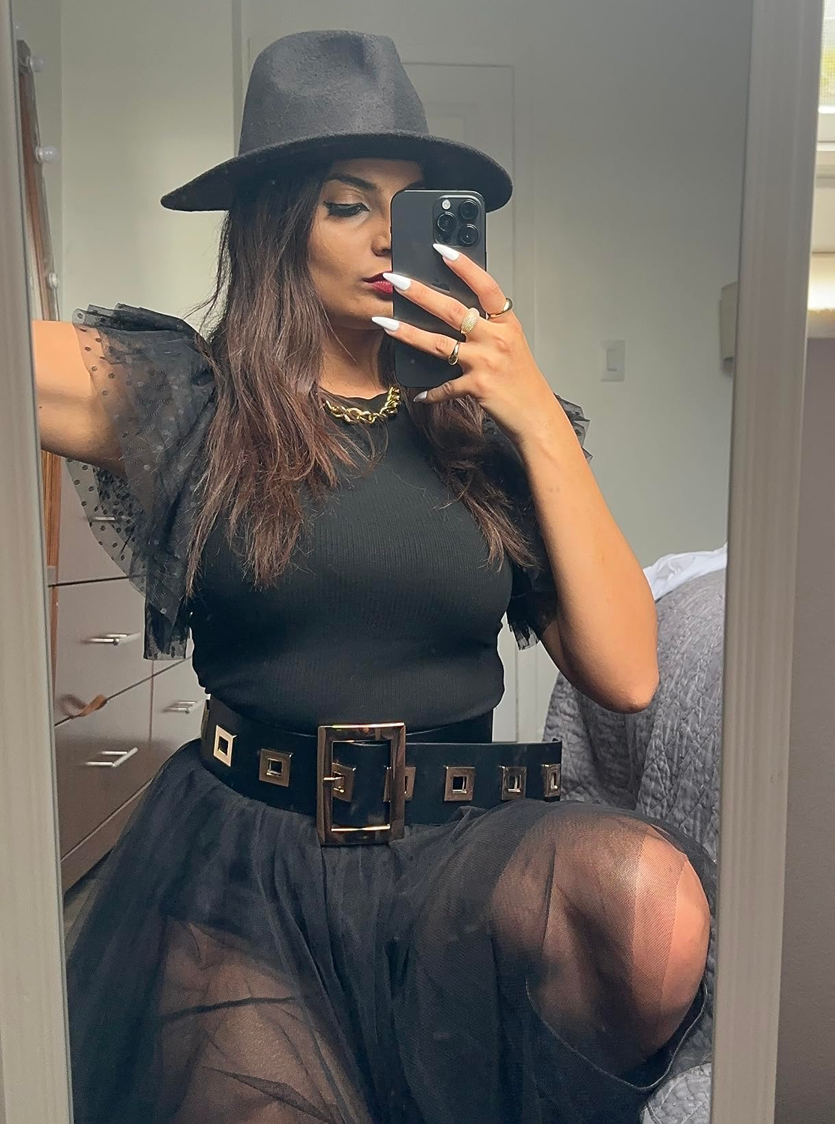 A reviewer in stylish black outfit with lace sleeves, wide belt, and fedora takes a mirror selfie