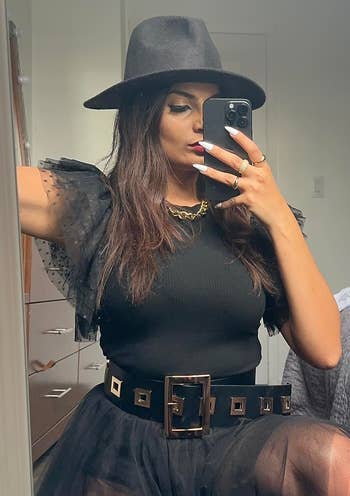 A reviewer in stylish black outfit with lace sleeves, wide belt, and fedora takes a mirror selfie