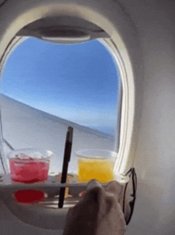 someone placing earbuds into holes on the bevledge, which is attached to a plane window and holding two drinks, a phone, and sunglasses