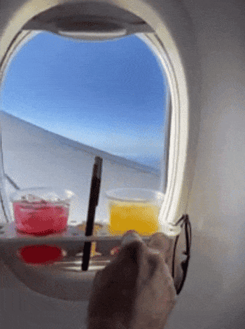 someone placing earbuds into holes on the bevledge, which is attached to a plane window and holding two drinks, a phone, and sunglasses