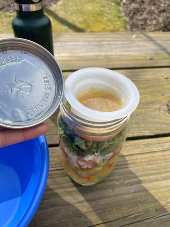 Salad dalam stoples dengan saus di tutupnya, diletakkan di atas meja piknik kayu dengan mangkuk biru dan botol air di dekatnya. Ideal untuk piknik