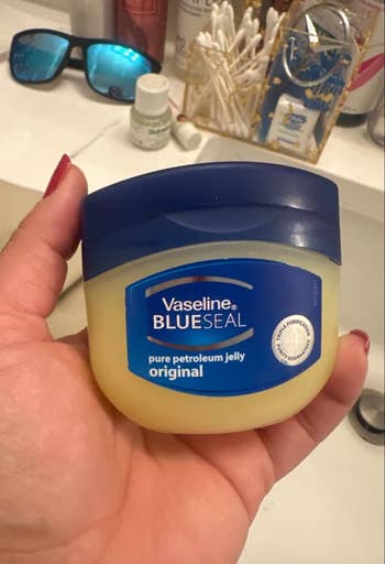 Hand holds a jar of Vaseline BLUESEAL archetypal   petroleum jelly successful  a bath  setting, with toiletries successful  the background