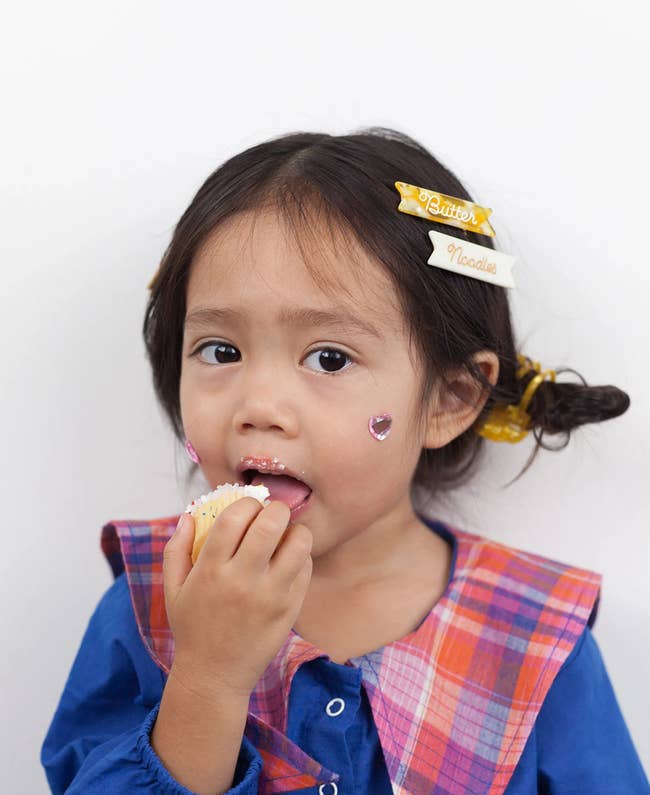 A child model with two clips in their hair: a yellow and white one that says butter and a white one that says noodles