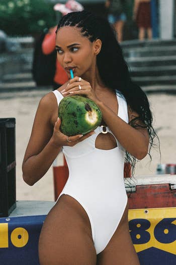 A person in a white one-piece swimsuit sips from a coconut on the beach
