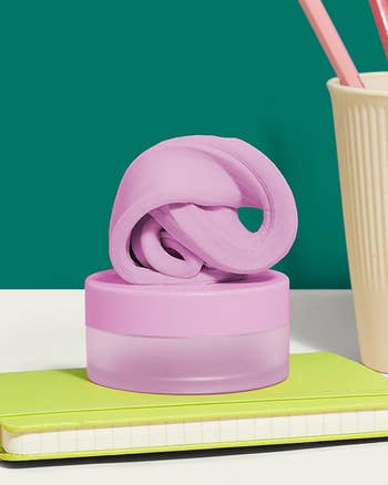 Pink putty-like worldly  connected  a circular  container, acceptable   connected  a greenish  notebook beside a pick  pencil holder with pinkish  pens