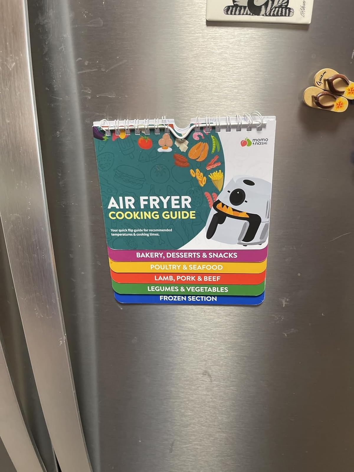 Air Fryer Cooking Guide hanging on a refrigerator, featuring categories for baking, poultry, meat, vegetables, and frozen foods
