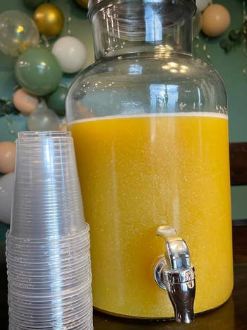 Large jar of orange juice with a spigot and stack of clear cups on a counter
