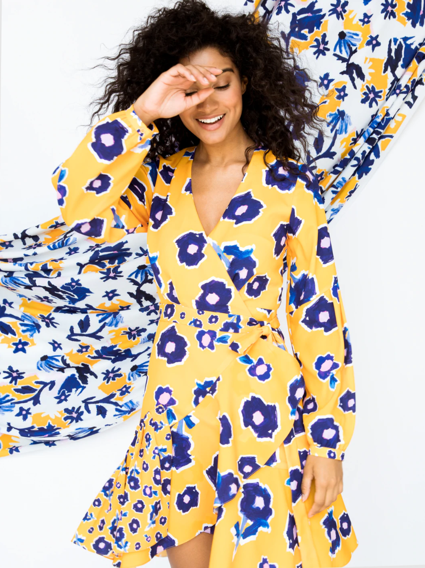 Model is wearing a bright yellow short wrap dress with blue flowers throughout