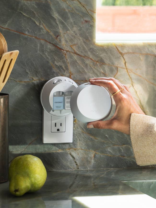 Person adjusting a sleek, modern air freshener plugged into a kitchen wall outlet, with a pear on the counter nearby