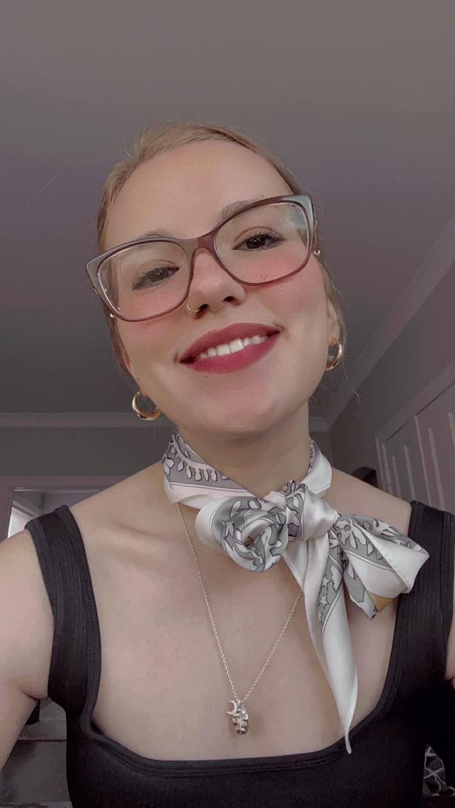 The reviewer smiling with glasses, scarf tied astir   neck, and a pendant necklace