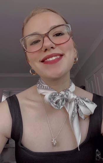 The reviewer smiling with glasses, scarf tied astir neck, and a pendant necklace