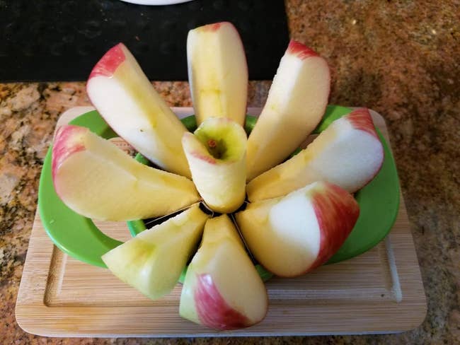 a reviewer photo of the sliced apple with red skin, divided using a green apple slicer, placed on a wooden cutting board