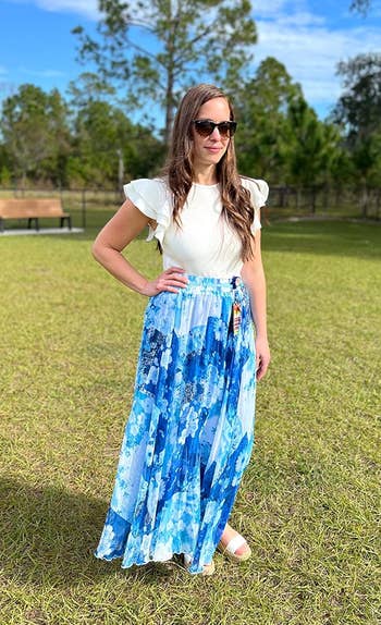 A Reviewer outdoors wearing a white ruffle-sleeve top, a flowing patterned skirt, and sunglasses