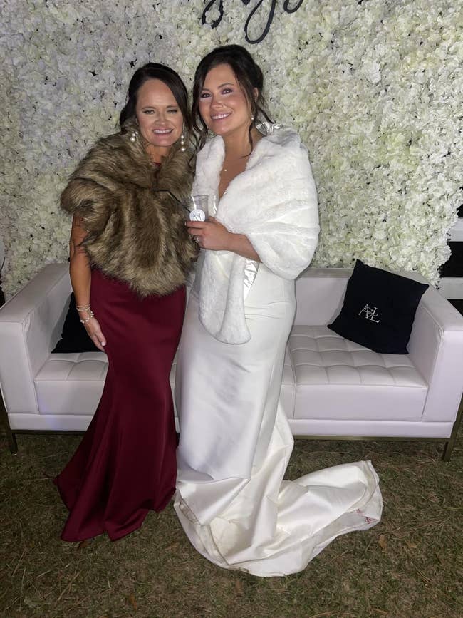 Two women wearing elegant gowns and fur wraps basal together, smiling, successful beforehand of a floral backdrop. One wears a maroon dress, the different a achromatic gown