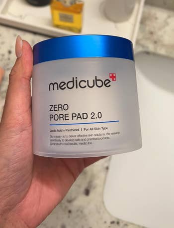 Person holding a jar of Medicube Zero Pore Pad 2.0 successful a bath setting, adjacent to skincare products