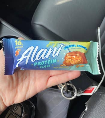 A hand holds an Alani Protein Bar in the Caramel Crunch flavor, with 16 grams of protein, in a car