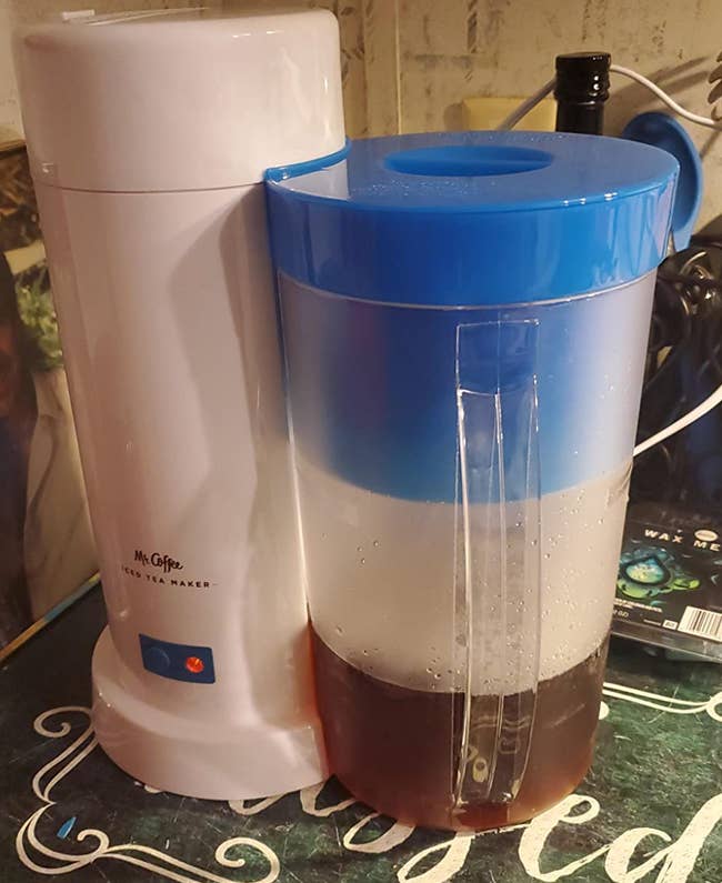 A cylinder shaped white iced tea maker with an attached pitcher full of iced tea 