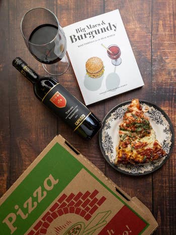 Pizza portion    connected  a plate, unfastened  vino  bottle, solid  of reddish  wine, pizza box, and wine-and-burger pairing publication  connected  a woody  table