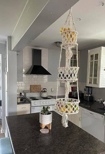 Macrame plant holder with yellow flowers hanging in a modern kitchen, featuring white cabinets, black countertop, and stainless steel appliances