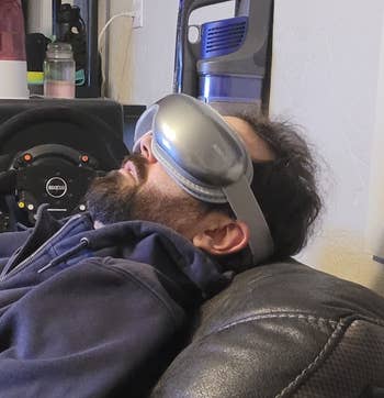 Person relaxing on a couch, wearing large, futuristic VR goggles, with a steering wheel visible in the background