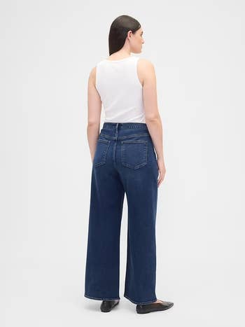 Person wearing wide-leg jeans and a white tank top, viewed from the back, standing against a plain backdrop