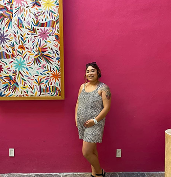 Lady in a patterned dress and sandals stands smiling in entrance of a purple wall with colourful artwork