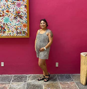 Woman in a patterned dress and sandals stands smiling in front of a pink wall with colorful artwork
