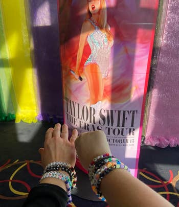 Person's hand with beaded bracelets reaching toward Taylor Swift's 'The Eras Tour' promo poster