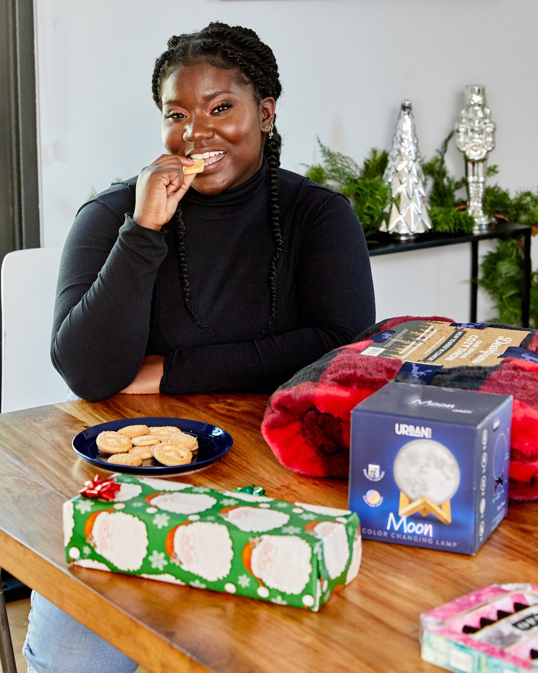 Elise biting into Christmas cookie at table with cozy blanket, wrapped gift, and moon color-changing lamp on it