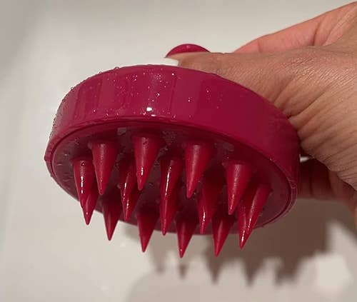 Person holding a purple scalp massager over a sink, indicating its use for hair care routines