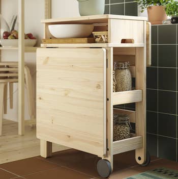 Wooden room  cart with a foldable tabletop, shelves holding jars of grains, and a tray with a vessel  connected  top; perfect  for compact kitchens