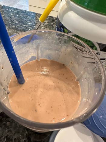 A blender with a smoothie mix and a straw on a kitchen counter, near protein powder containers