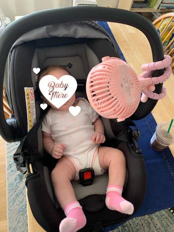 Baby sitting in a car seat with a fan attached, wearing a onesie and pink socks. Starbucks drink on the table in the background