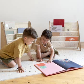 Two children sit on a floor, engaging with a large picture book. Behind them, books are displayed on low wooden shelves