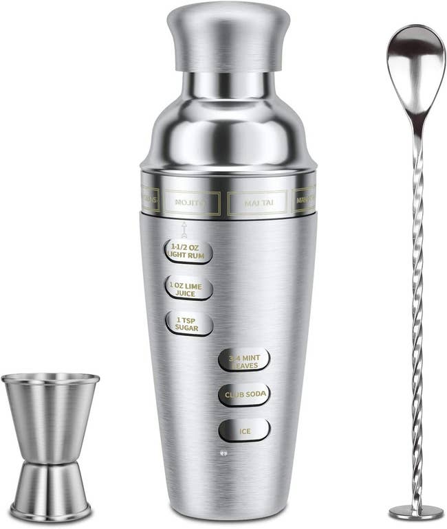 A stainless steel cocktail shaker with cut outs on the side showing ingredients for different drinks, along with a with a jigger and mixing spoon 